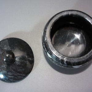 Pu&ograve; includere: Un vaso di marmo nero con coperchio abbinato. Il vaso &egrave; rotondo con un'ampia apertura e il coperchio ha un piccolo pomello. Il vaso e il coperchio hanno una superficie lucida e riflettente e il marmo presenta un motivo a vortice.