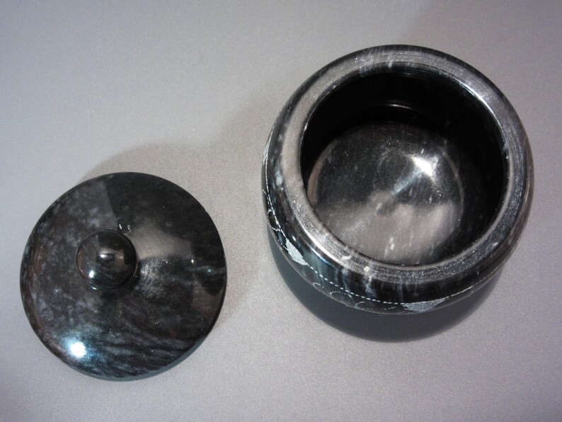 Pu&ograve; includere: Un vaso di marmo nero con coperchio abbinato. Il vaso &egrave; rotondo con un'ampia apertura e il coperchio ha un piccolo pomello. Il vaso e il coperchio hanno una superficie lucida e riflettente e il marmo presenta un motivo a vortice.