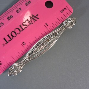 May include: A silver filigree brooch with a row of clear stones set in the center. The brooch is about 1 inch long and is measured against a pink ruler.