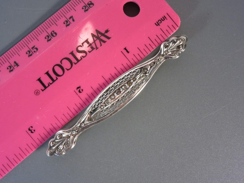May include: A silver filigree brooch with a row of clear stones set in the center. The brooch is about 1 inch long and is measured against a pink ruler.