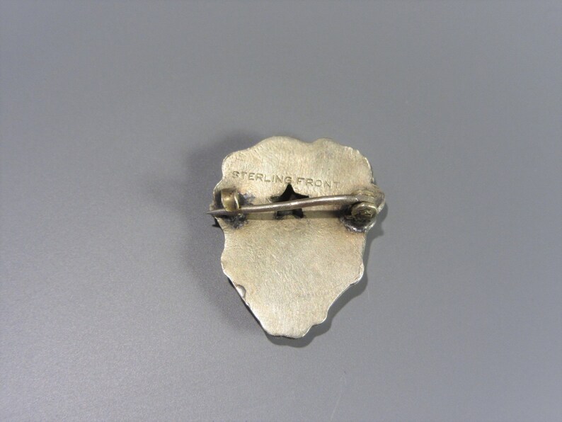 Pode incluir: Pin de tom prateado com um design detalhado. A parte de tr&aacute;s do pin tem as palavras "STERLING FRONT" gravadas.