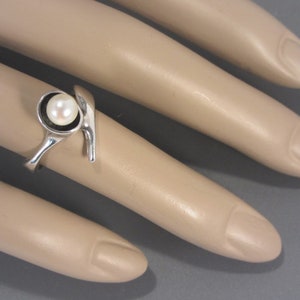 Puede incluir: Un anillo de plata con una sola perla blanca engastada en un bisel circular. El anillo tiene un dise&ntilde;o &uacute;nico y abstracto con una banda curva.