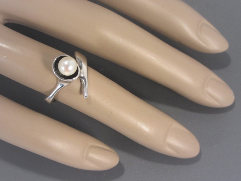 Puede incluir: Un anillo de plata con una sola perla blanca engastada en un bisel circular. El anillo tiene un dise&ntilde;o &uacute;nico y abstracto con una banda curva.