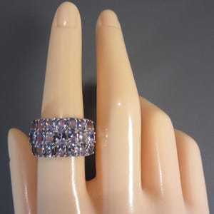 May include: A wide, silver-toned ring featuring three rows of oval-cut, light purple gemstones. The ring is displayed on a mannequin hand against a gray background. The gemstones are closely set, creating a continuous band of color.