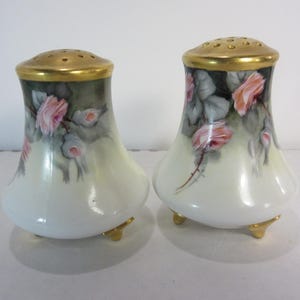 May include: A pair of antique salt and pepper shakers. The shakers are white with a gold rim and gold feet. They are decorated with pink roses and green leaves. The shakers are approximately 4 inches tall.