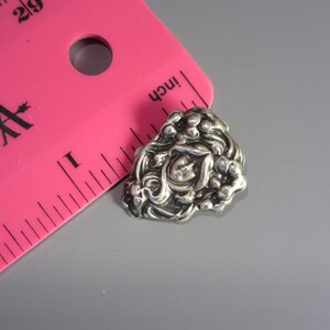 Pode incluir: Um broche de metal prateado com um design floral. O broche apresenta uma flor central com um cora&ccedil;&atilde;o em relevo no centro, rodeado por flores e folhas menores.
