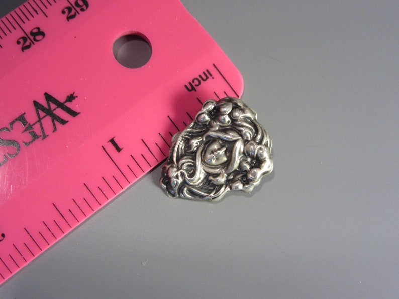 Pode incluir: Um broche de metal prateado com um design floral. O broche apresenta uma flor central com um cora&ccedil;&atilde;o em relevo no centro, rodeado por flores e folhas menores.