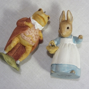 May include: Two ceramic figurines featuring a frog dressed in a brown coat and patterned vest, and a rabbit wearing a blue dress with a white apron and holding a small woven basket. The rabbit figurine has the text Beatrix Potter printed on the base.