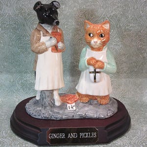 May include: A ceramic figurine featuring a black dog and an orange cat standing on a gray stone base. Both animals wear white aprons. The dog holds a brown bag, and the cat holds a white package with a black cross. A plaque reads GINGER AND PICKLES.