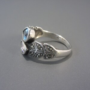 May include: A silver ring featuring three stones. The stones are light blue, light pink, and clear. The ring is adorned with a design of small, dark grey stones.