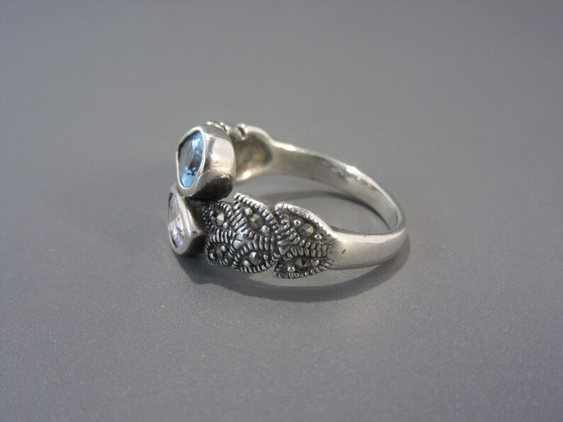 May include: A silver ring featuring three stones. The stones are light blue, light pink, and clear. The ring is adorned with a design of small, dark grey stones.