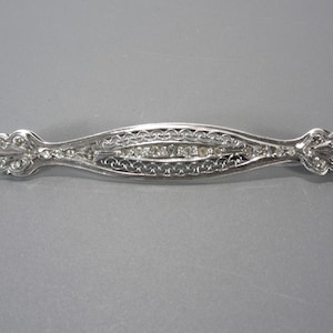 May include: A silver brooch with a delicate filigree design and a row of clear stones in the center.