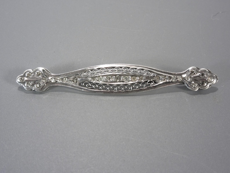 May include: A silver brooch with a delicate filigree design and a row of clear stones in the center.