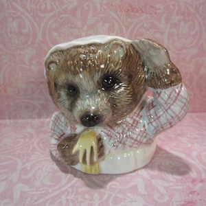 May include: A ceramic bear figurine featuring a brown bear wearing a white nightcap and a white nightshirt with a red plaid pattern. The bear is depicted with one paw raised to its head and a yellow cloth draped over its other paw in a seated pose.