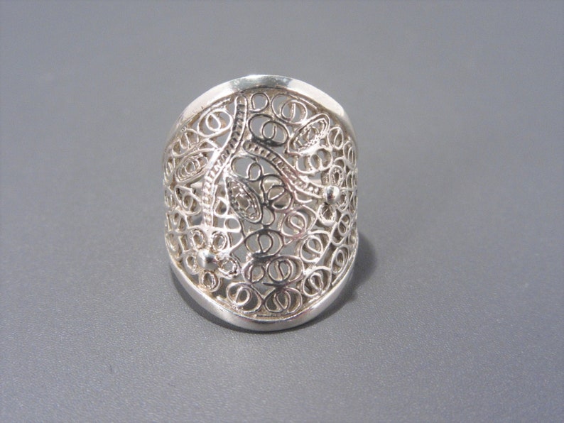 May include: A silver filigree ring with a delicate, intricate design. The ring is wide and has a rounded shape.