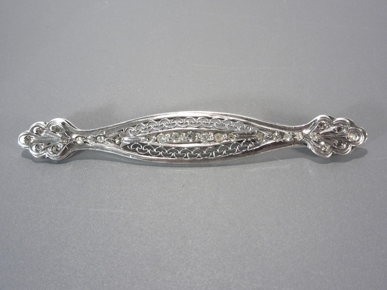 May include: A silver filigree brooch with a row of clear rhinestones in the center.