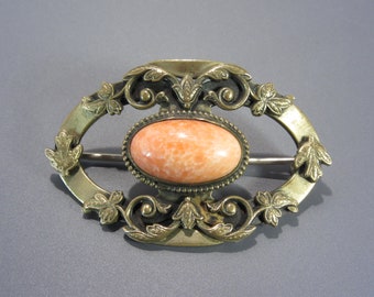 Può includere: Un fermaglio dorato con una pietra ovale di colore pesca incastonata in una cornice decorativa. La cornice presenta intricati disegni floreali.