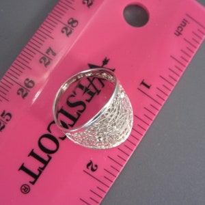 May include: A silver ring with a delicate, intricate design. The ring is measured against a pink ruler, showing it is slightly over 2.5 cm in diameter.