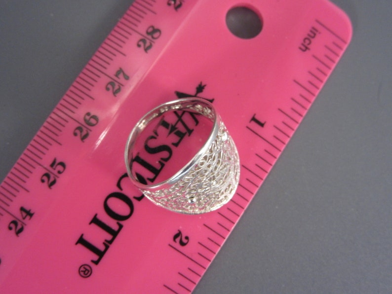 May include: A silver ring with a delicate, intricate design. The ring is measured against a pink ruler, showing it is slightly over 2.5 cm in diameter.