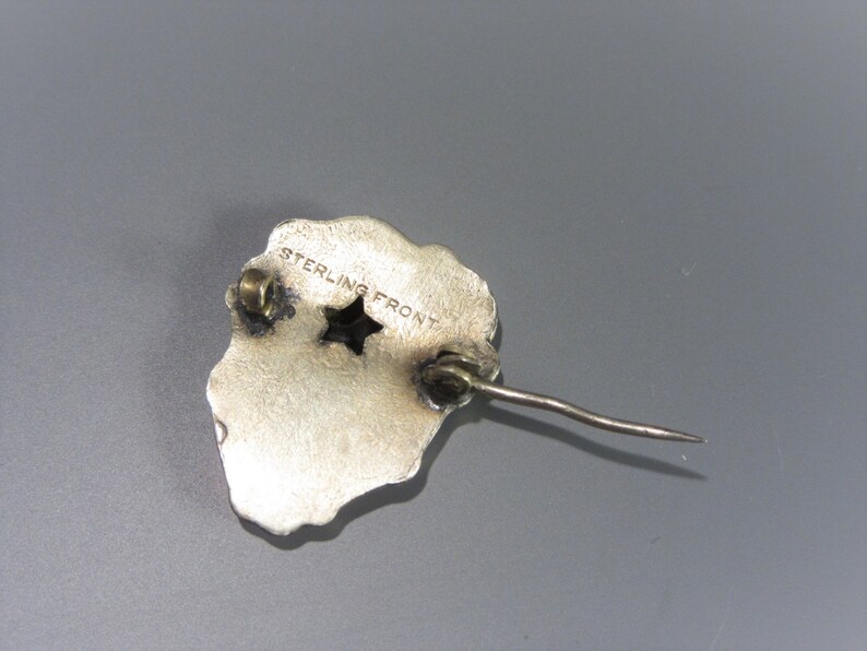 Pode incluir: Broche prateada com um design em forma de estrela e as palavras "STERLING FRONT" gravadas no verso.