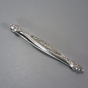 May include: Silver sugar tongs with an ornate design. The tongs are closed and have a decorative, swirling pattern.
