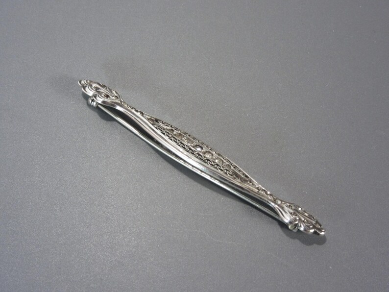 May include: Silver sugar tongs with an ornate design. The tongs are closed and have a decorative, swirling pattern.