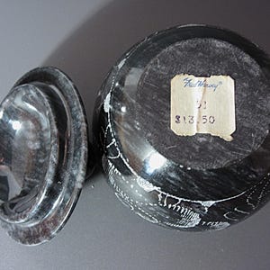 Pu&ograve; includere: Un vaso in pietra scolpita nera e grigia con coperchio. Il vaso presenta disegni incisi bianchi e un'etichetta con la scritta "Fred Harvey". Il coperchio &egrave; di forma ovale e presenta un motivo a spirale. Il vaso &egrave; un oggetto decorativo.