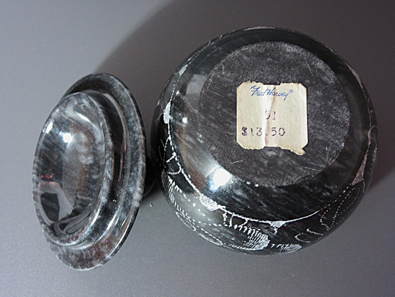 Pu&ograve; includere: Un vaso in pietra scolpita nera e grigia con coperchio. Il vaso presenta disegni incisi bianchi e un'etichetta con la scritta "Fred Harvey". Il coperchio &egrave; di forma ovale e presenta un motivo a spirale. Il vaso &egrave; un oggetto decorativo.