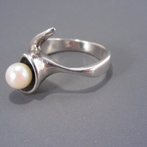 Puede incluir: Un anillo de plata con una perla engastada en un dise&ntilde;o &uacute;nico y abstracto. El anillo est&aacute; hecho de metal liso y pulido y presenta una forma curva y org&aacute;nica.