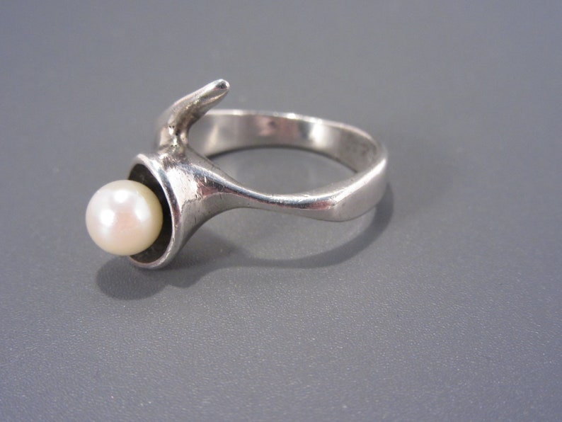 Puede incluir: Un anillo de plata con una perla engastada en un dise&ntilde;o &uacute;nico y abstracto. El anillo est&aacute; hecho de metal liso y pulido y presenta una forma curva y org&aacute;nica.