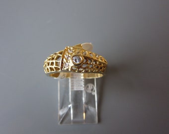 Può includere: Un anello d'oro con un design delicato e traforato e una singola pietra preziosa trasparente al centro. L'anello è incastonato con pietre preziose più piccole intorno alla fascia.