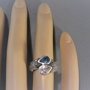 May include: A silver ring with a delicate filigree design features two pear-shaped gemstones. The larger gemstone is a light blue colour, while the smaller gemstone is a pale pink.