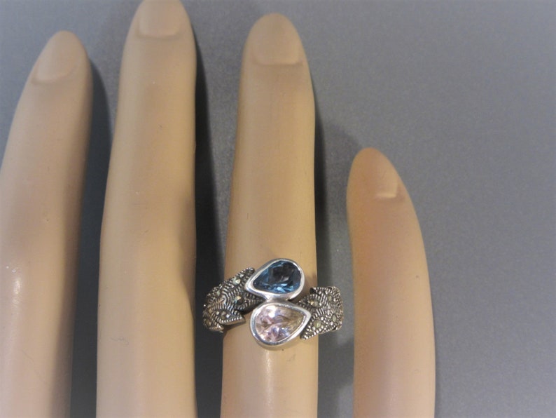 May include: A silver ring with a delicate filigree design features two pear-shaped gemstones. The larger gemstone is a light blue colour, while the smaller gemstone is a pale pink.