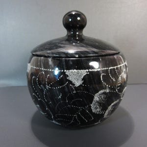 Pu&ograve; includere: Un vaso di marmo nero con coperchio e una piccola maniglia rotonda. Il vaso ha una forma arrotondata ed &egrave; decorato con motivi floreali bianchi incisi. Il coperchio si adatta perfettamente. &Egrave; un contenitore decorativo.