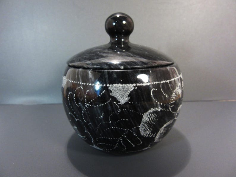 Pu&ograve; includere: Un vaso di marmo nero con coperchio e una piccola maniglia rotonda. Il vaso ha una forma arrotondata ed &egrave; decorato con motivi floreali bianchi incisi. Il coperchio si adatta perfettamente. &Egrave; un contenitore decorativo.
