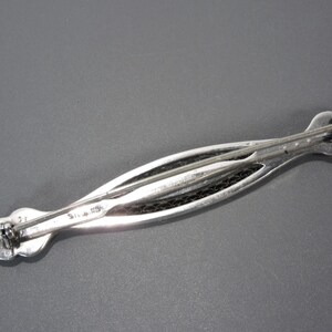 May include: A silver brooch with a delicate, intricate design. The brooch is shaped like a long, narrow leaf with a textured surface. The back of the brooch has a pin and catch closure.