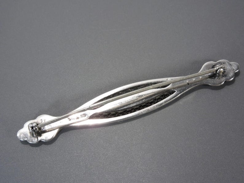 May include: A silver brooch with a delicate, intricate design. The brooch is shaped like a long, narrow leaf with a textured surface. The back of the brooch has a pin and catch closure.