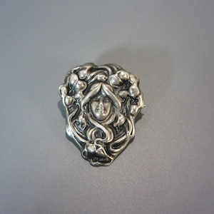 Pode incluir: Um broche de prata com um design detalhado de um rosto de mulher rodeado de flores.