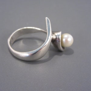 Puede incluir: Un anillo de plata con un dise&ntilde;o curvo y una perla blanca en el centro.