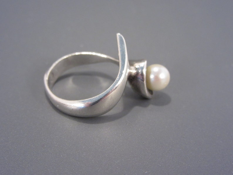 Puede incluir: Un anillo de plata con un dise&ntilde;o curvo y una perla blanca en el centro.