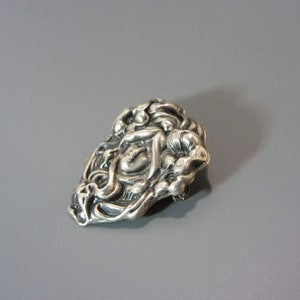 Pode incluir: Um broche de prata com um design detalhado de um rosto de mulher rodeado de flores.
