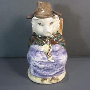 May include: A ceramic character jug shaped like a pig wearing a brown hat, a dark green shawl, and a purple dress. The pig figure has white fur, pointed ears, and is holding a yellow object in its hands. The piece features a glossy finish.