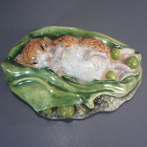 May include: A ceramic figurine of a small, brown and white mouse sleeping inside a green pea pod. The mouse is curled up with its eyes closed, surrounded by several small, round, green peas nestled within the textured, glossy green pod leaves.