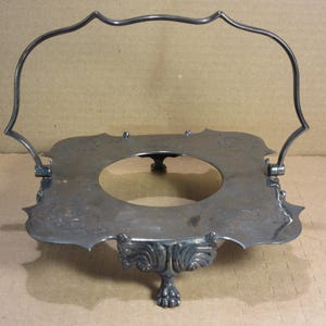May include: A silver-toned metal serving tray with a decorative handle. The tray has a square shape with a circular opening in the center and ornate detailing around the edges. The tray is supported by three decorative feet.