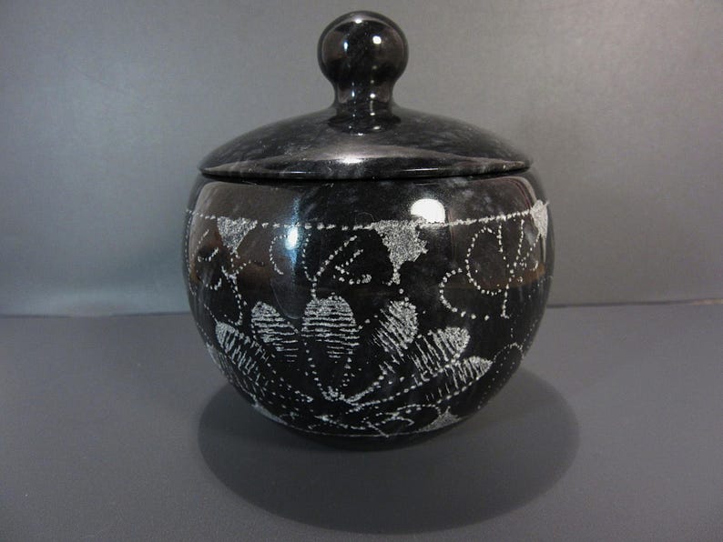 Pu&ograve; includere: Un vaso di marmo nero con coperchio. Il vaso rotondo presenta un motivo floreale bianco. Il coperchio ha una piccola maniglia rotonda. Questo vaso &egrave; probabilmente per la conservazione o l'esposizione.