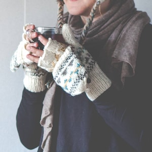 May include: A person wearing a black long-sleeve shirt, holding a clear glass of tea. The person is wearing knitted fingerless gloves with a geometric pattern in cream, blue, and gray. A brown scarf and hat complete the cozy look.