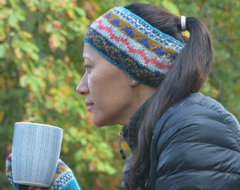May include: A woman wearing a knitted headband and mittens, holding a light blue mug. The headband and mittens feature a colorful geometric pattern with blue, red, and yellow accents. She is wearing a black jacket.