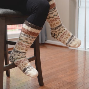 May include: A pair of knee-high socks with a patterned design in cream, brown, and muted green. The socks have a ribbed cuff and a detailed geometric pattern. The socks are worn by someone sitting on a wooden stool.