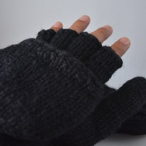 May include: A pair of dark gray fingerless gloves, knitted with a textured yarn. The gloves have open fingertips and a wrist cuff. The image shows the gloves on a hand against a light gray background. The gloves appear to be warm and suitable for cold weather.