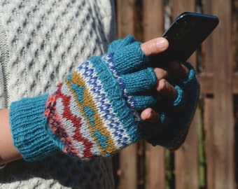 May include: Teal knitted fingerless gloves with a colourful geometric pattern in red, white, yellow, and blue. The gloves are being held by a person wearing a white cable-knit sweater, and holding a black smartphone.